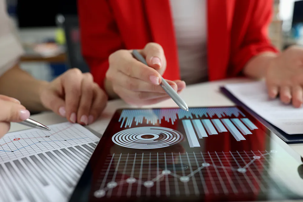A person in a red blazer pointing at a tablet displaying data charts and graphs during a business meeting, with printed reports and a pen visible on the table.