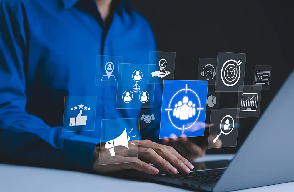 A person in a blue shirt typing on a laptop, surrounded by floating digital icons representing marketing and sales concepts including audience targeting, location, customer ratings, megaphone, analytics, advertising, and customer support.