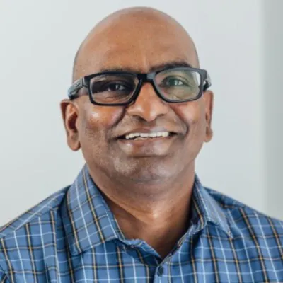 Headshot of a smiling man wearing glasses and a blue plaid shirt against a light gray background.